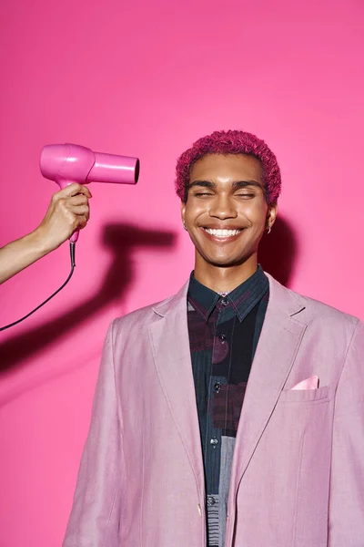 Homem alegre agindo como boneca masculina em traje vibrante posando em pano de fundo rosa, mão com secador de cabelo — Fotografia de Stock
