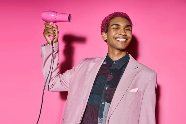 Good looking stylish man in bold attire smiling weirdly with closed eyes and hairdryer in hands — Stock Photo
