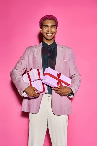 Jolly african american man smiling weirdly at camera and holding presents in hands on pink backdrop — Stock Photo