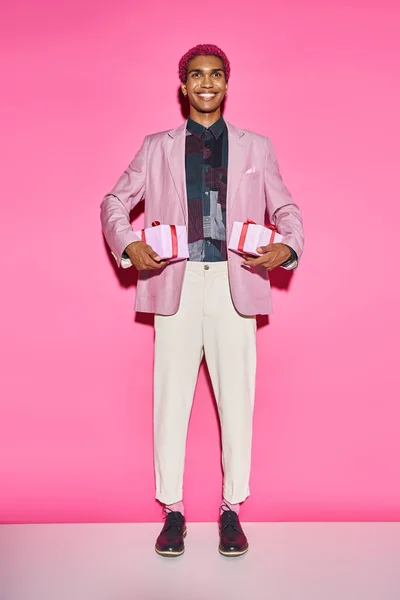Gut aussehender Mann mit lockigem Haar posiert unnatürlich und lächelt mit Geschenken auf rosa Hintergrund — Stockfoto