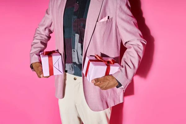 Vue recadrée du jeune homme en tenue élégante posant avec des cadeaux dans les deux mains sur fond rose — Photo de stock