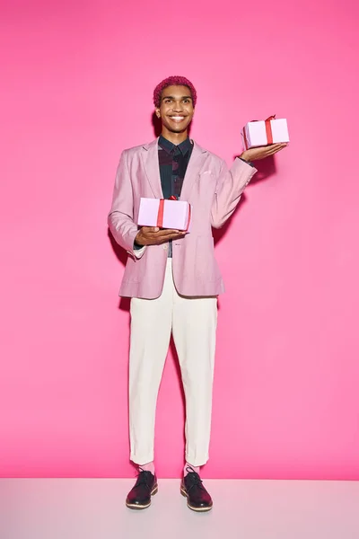 Bel homme afro-américain en tenue vibrante posant avec des cadeaux dans les mains sur fond rose — Photo de stock