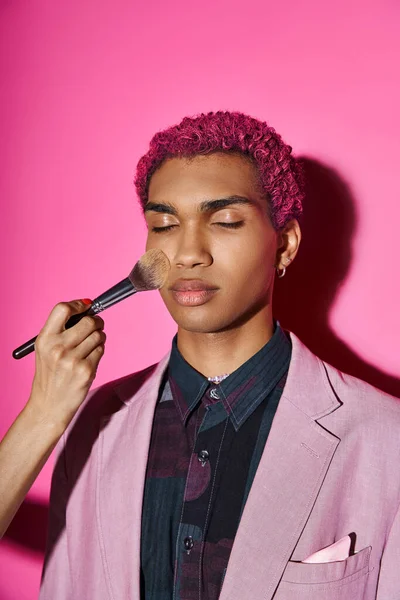 Homem bonito com cabelo rosa encaracolado posando com olhos fechados, mão com escova de maquiagem, boneca como — Fotografia de Stock