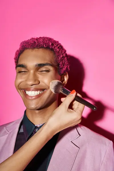Веселий молодий чоловік, який щиро посміхається закритими очима, рука з пензлем для макіяжу на рожевому тлі — стокове фото