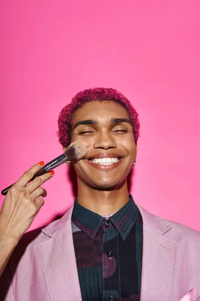 Modelo masculino elegante alegre em traje vívido sorrindo com olhos fechados, mão com escova de maquiagem — Fotografia de Stock