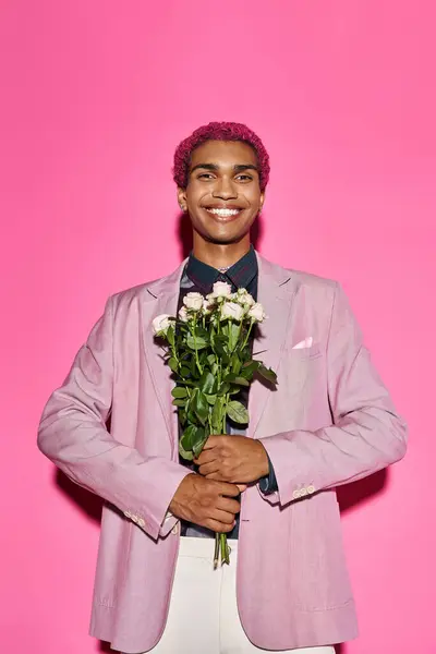 Fröhlicher Mann mit lockigem Haar in rosa Blazer posiert mit Rosenstrauß und blickt in die Kamera — Stockfoto