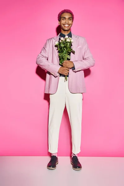 Homem bonito com cabelo encaracolado posando de forma não natural e sorrindo com buquê de rosa na frente dele — Fotografia de Stock