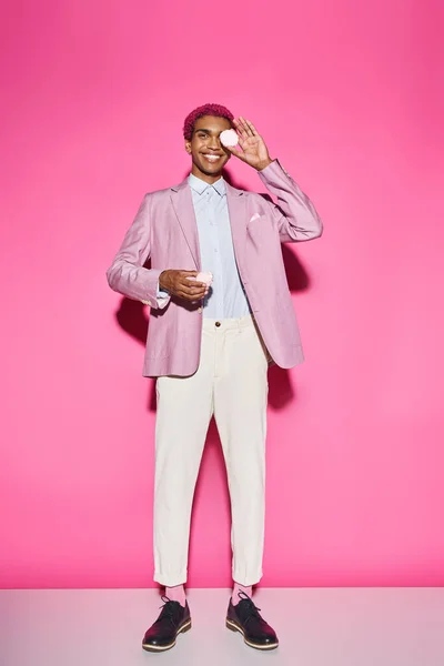 Hombre guapo sonriendo antinaturalmente y de pie quieto con zefir en las manos sobre el telón de fondo rosa - foto de stock