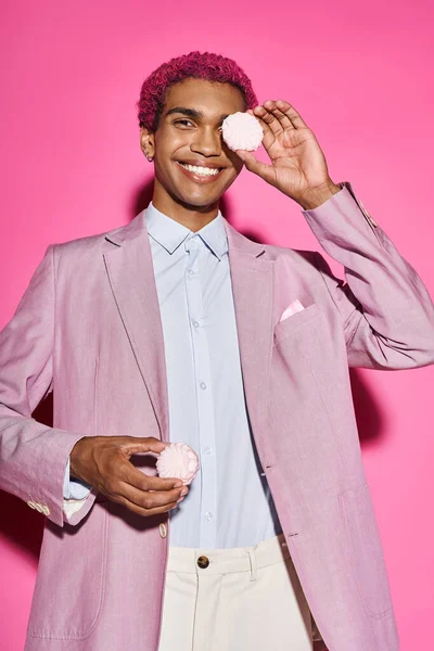 Joven hombre elegante sonriendo antinatural y posando con zefir en sus manos posando sobre telón de fondo rosa - foto de stock