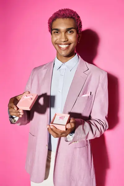 Homem bonito sorrindo para a câmera de forma não natural e mostrando pequeno presente rosa no fundo rosa — Fotografia de Stock