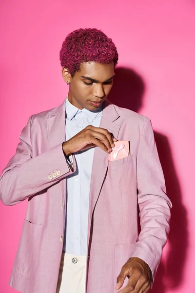Bel homme aux cheveux rose bouclés en tenue élégante sur fond rose avec présent dans sa poche — Photo de stock