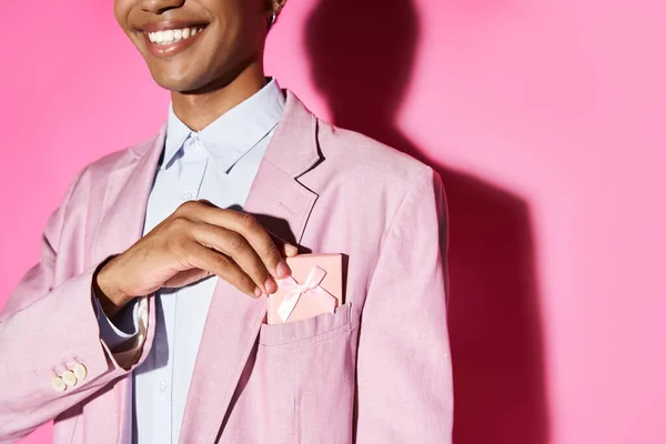 Ausgeschnittene Ansicht eines Smileys junger Mann in rosa Blazer, der Geschenk in seine Tasche auf rosa Hintergrund legt — Stockfoto