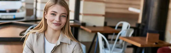Happy young blonde woman in trench coat looking at camera and sitting outdoors in cafe, banner — Stock Photo
