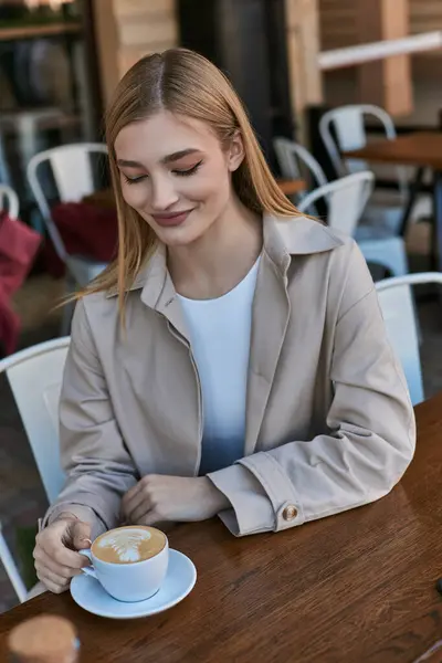 Fröhliche junge blonde Frau im Trenchcoat genießt ihre Tasse Kaffee im Freien im Café, Cappuccino — Stockfoto