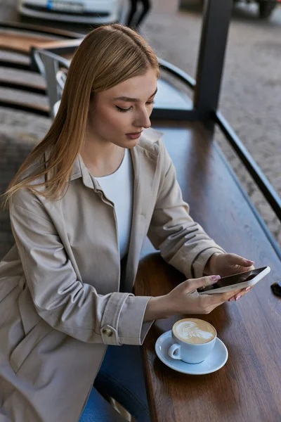 Blonde young woman in trench coat using smartphone near cup with cappuccino in outdoor cafe — Stock Photo