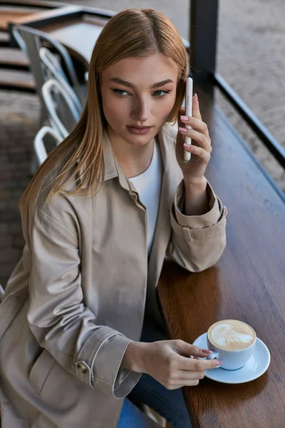 Blonde junge Frau im Trenchcoat mit Smartphone in der Nähe von Tasse mit Cappuccino im Outdoor-Café — Stockfoto