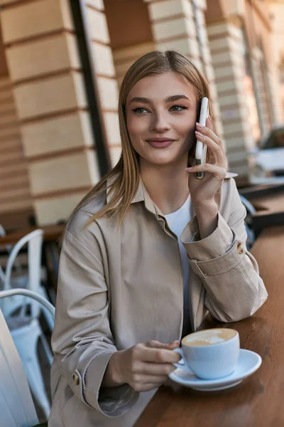 Felice giovane donna in trench avendo chiamata su smartphone vicino tazza con cappuccino in caffè all'aperto — Foto stock