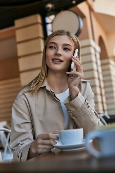 Jovem alegre em casaco de trincheira ter chamada no smartphone perto da xícara com cappuccino no café ao ar livre — Fotografia de Stock