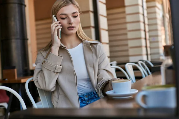 Hübsche junge Frau im Trenchcoat mit Smartphone in der Nähe von Tasse mit Cappuccino im Outdoor-Café — Stockfoto