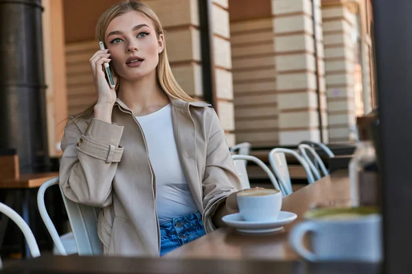 Hübsche Frau im Trenchcoat mit Smartphone in der Nähe von Tasse mit Cappuccino im Outdoor-Café — Stockfoto