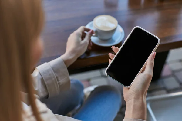 Ritagliato giovane donna in trench utilizzando smartphone vicino a tazza di caffè mentre seduto in caffè all'aperto — Foto stock
