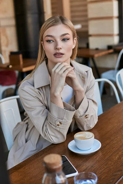 Dreamy young blonde woman in trench coat sitting near cup of coffee and smartphone outdoors in cafe — Stock Photo