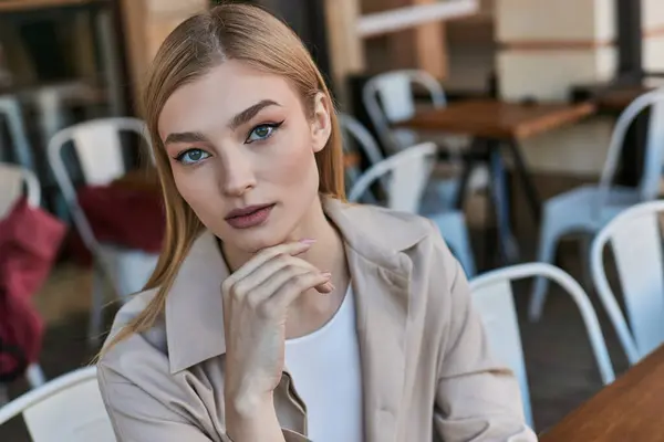 Porträt einer hübschen blonden Frau im Trenchcoat, die in die Kamera blickt, während sie draußen im Café sitzt — Stockfoto