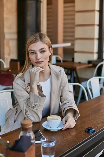 Junge blonde Frau im Trenchcoat genießt ihre Tasse Cappuccino im Freien im Café, am Wochenendmorgen — Stockfoto