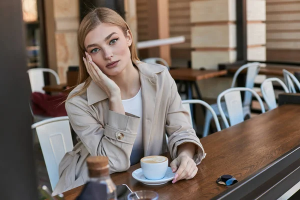 Attraktive junge Frau im Trenchcoat genießt ihre Tasse Cappuccino im Freien im Café, Wochenende — Stockfoto
