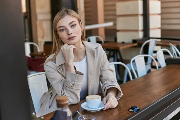 Verträumte blonde Frau im Trenchcoat genießt ihre Tasse Cappuccino im Freien im Café, Wochenendstimmung — Stockfoto