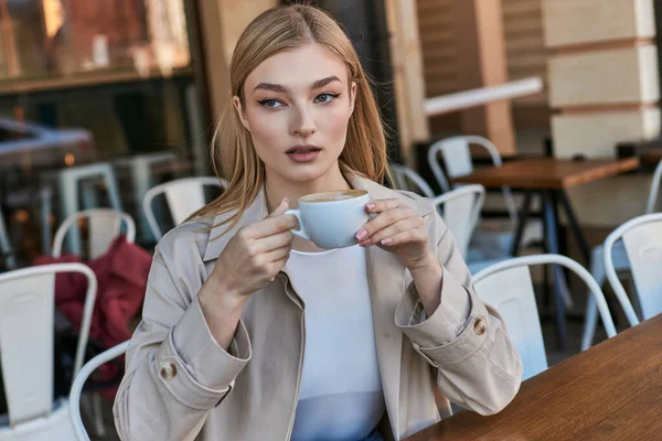 Verträumte blonde Frau im Trenchcoat genießt ihre Tasse Cappuccino, während sie draußen im Café sitzt — Stockfoto