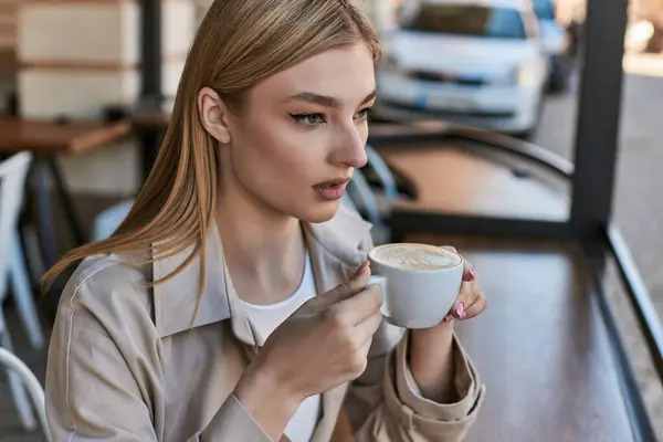 Verträumte junge Frau im Trenchcoat genießt ihre Tasse Cappuccino, während sie draußen im Café sitzt — Stockfoto