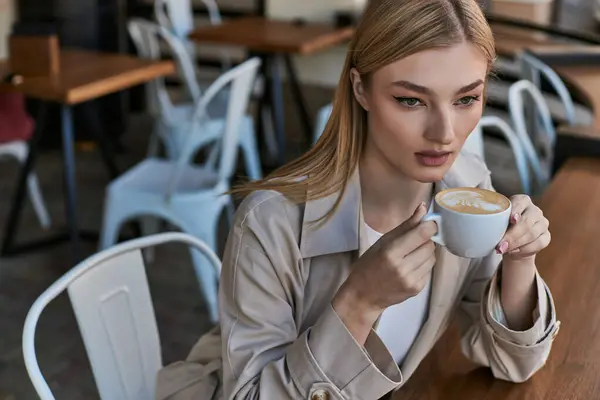 Verträumte hübsche Frau im Trenchcoat genießt ihre Tasse Cappuccino, während sie draußen im Café sitzt — Stockfoto