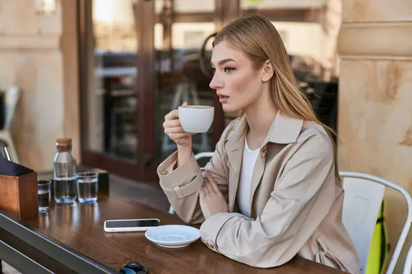 Verträumte junge Frau im Trenchcoat genießt eine Tasse Cappuccino, während sie neben dem Smartphone im Café sitzt — Stockfoto