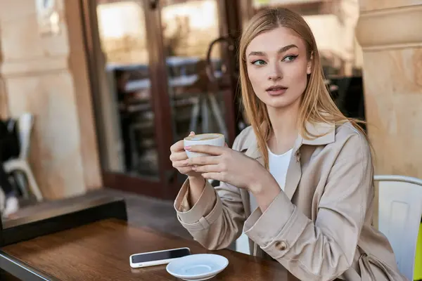 Hübsche junge Frau im Trenchcoat genießt eine Tasse Cappuccino, während sie neben dem Smartphone im Café sitzt — Stockfoto