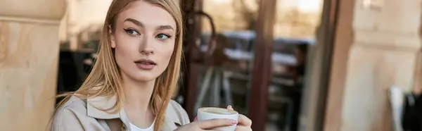 Мечтательная и молодая женщина в плаще наслаждается чашкой капучино, сидя в кафе, баннер — стоковое фото