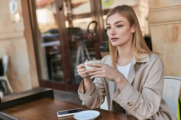 Hübsche junge Frau im Trenchcoat trinkt Cappuccino, während sie neben dem Smartphone im Café sitzt — Stockfoto