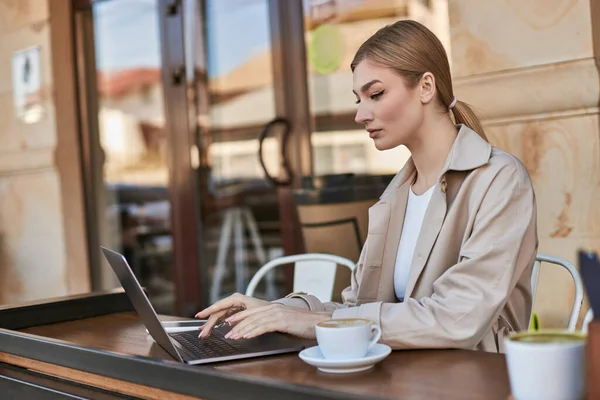 Jeune femme blonde tapant sur ordinateur portable tout en travaillant à distance près de tasse de café dans le café à l'extérieur — Photo de stock