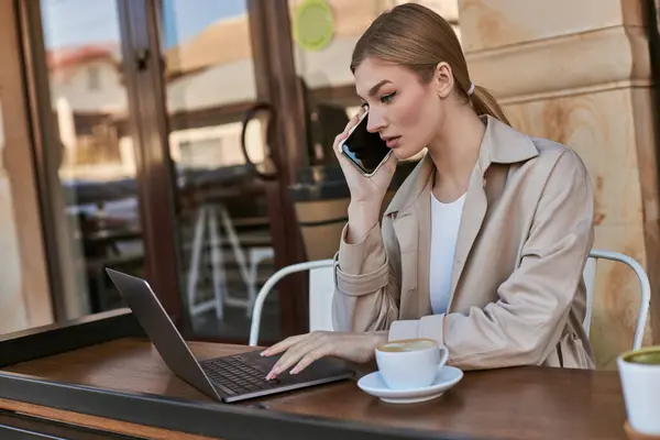 Young blonde freelancer typing on laptop while talking on smartphone and working remotely in cafe — Stock Photo