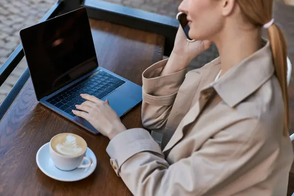 Blonde freelancer typing on laptop while talking on smartphone and working remotely in cafe — Stock Photo
