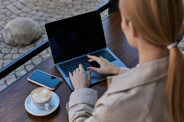 Young blonde freelancer typing on laptop while working remotely near cup of coffee in cafe outdoors — Stock Photo