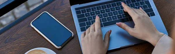Top view of woman typing on laptop while working near cup of coffee and smartphone in cafe, banner — Stock Photo