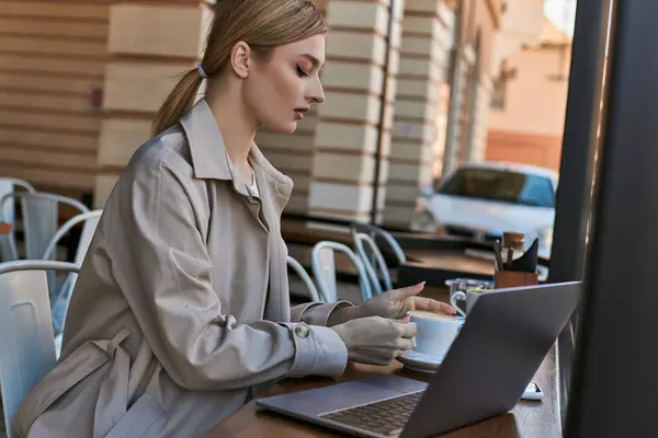 Young blonde woman taking cup of coffee near laptop on table in cafe outdoors, remote work — Stock Photo