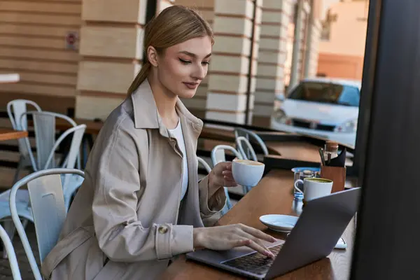 Young blonde woman holding cup of coffee and using laptop on table in cafe outdoors, remote work — Stock Photo