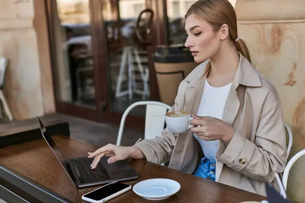 Blonde woman holding cup of coffee and using laptop on table in cafe outdoors, remote work — Stock Photo