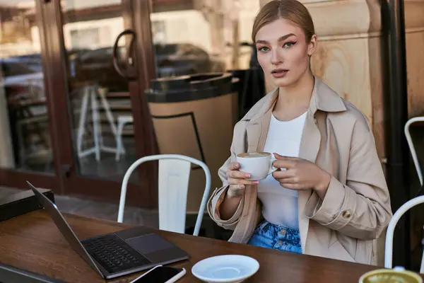 Atractiva mujer sosteniendo la taza de café y el uso de la computadora portátil en la mesa en la cafetería al aire libre, el trabajo remoto - foto de stock
