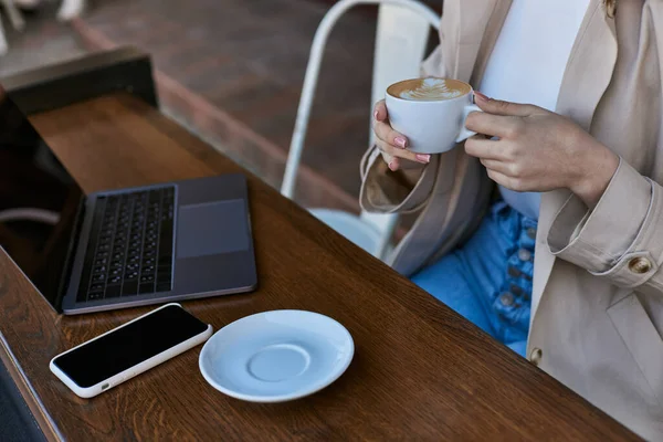 Cropped view of woman holding cup of coffee near laptop on table in cafe outdoors, remote work — Stock Photo