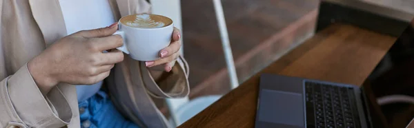 Cropped banner of woman holding cup of coffee near laptop on table in cafe outdoors, remote work — Stock Photo