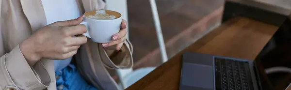 Cropped banner of woman holding cup of coffee near laptop on table in cafe outdoors, remote work — Stock Photo