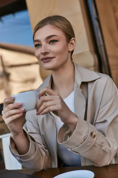 Blonde junge Frau im Trenchcoat genießt ihre Tasse Cappuccino im Café, entspannte Atmosphäre — Stockfoto